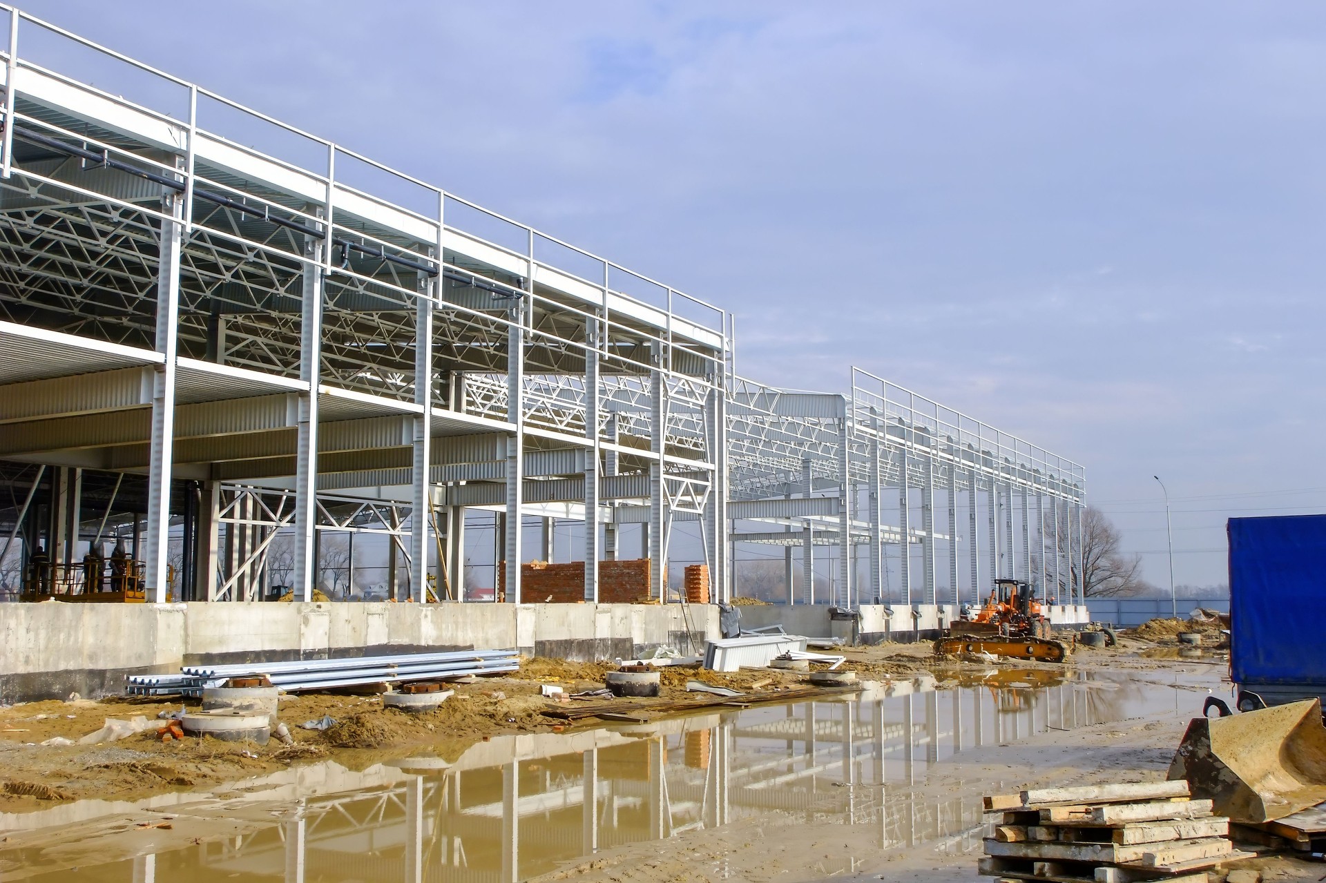 A large industrial building with a steel framework is under construction on a muddy site.